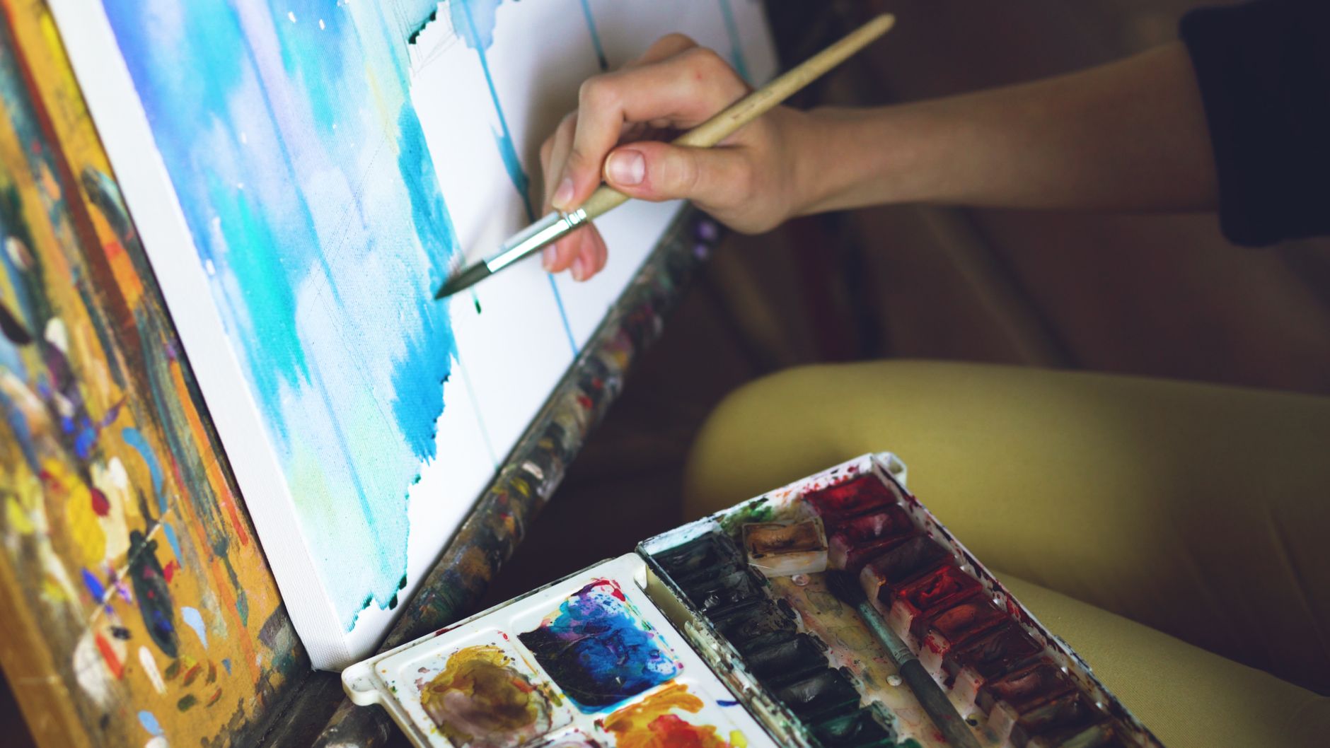 Close-up of an artist's hand painting on a canvas using watercolors with a palette.