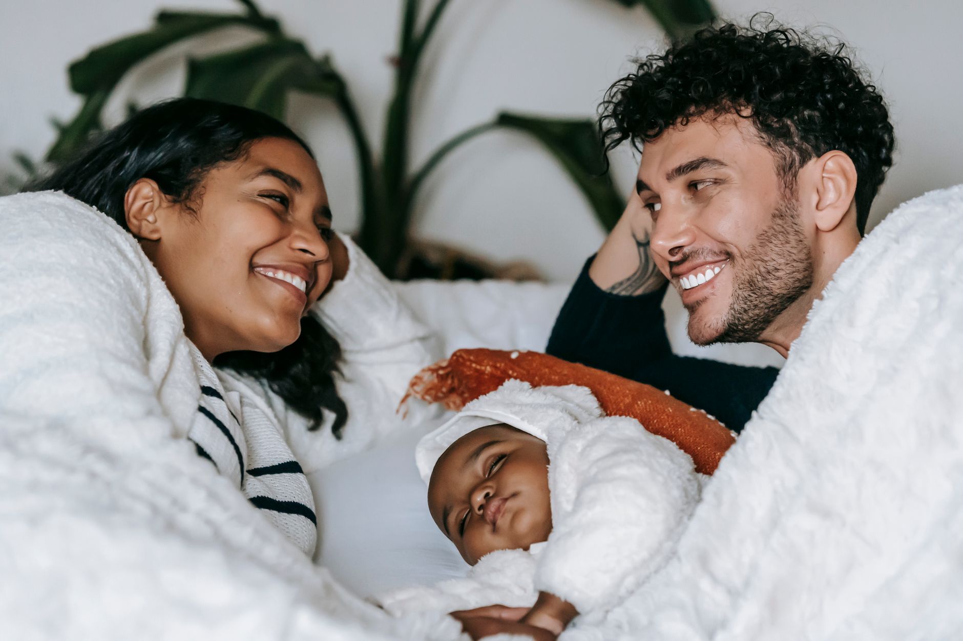 Positive ethnic couple in casual clothes lying near baby in comfortable bed and looking at each other