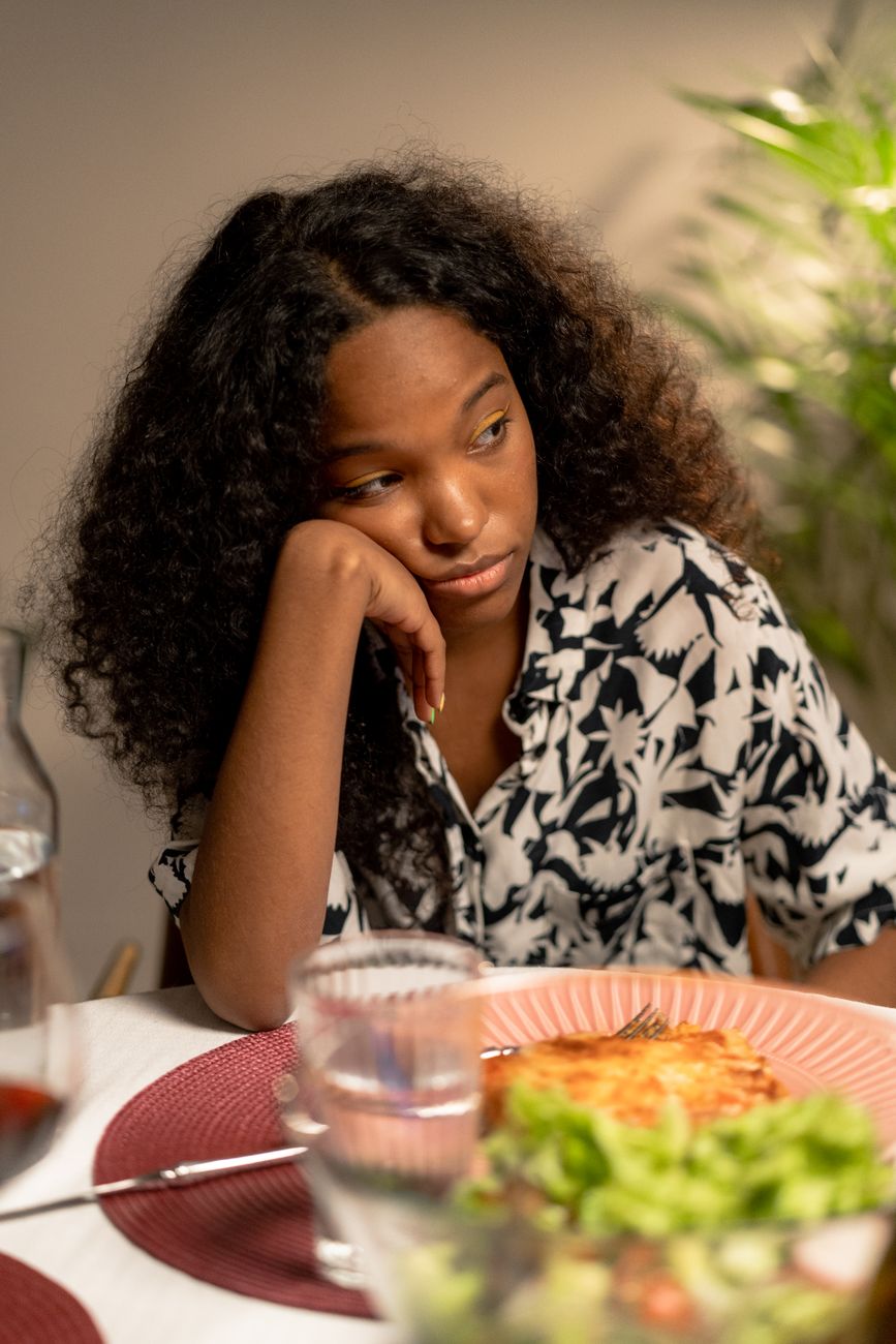 Teen girl with curly hair sits pensively at dinner table with a meal.
